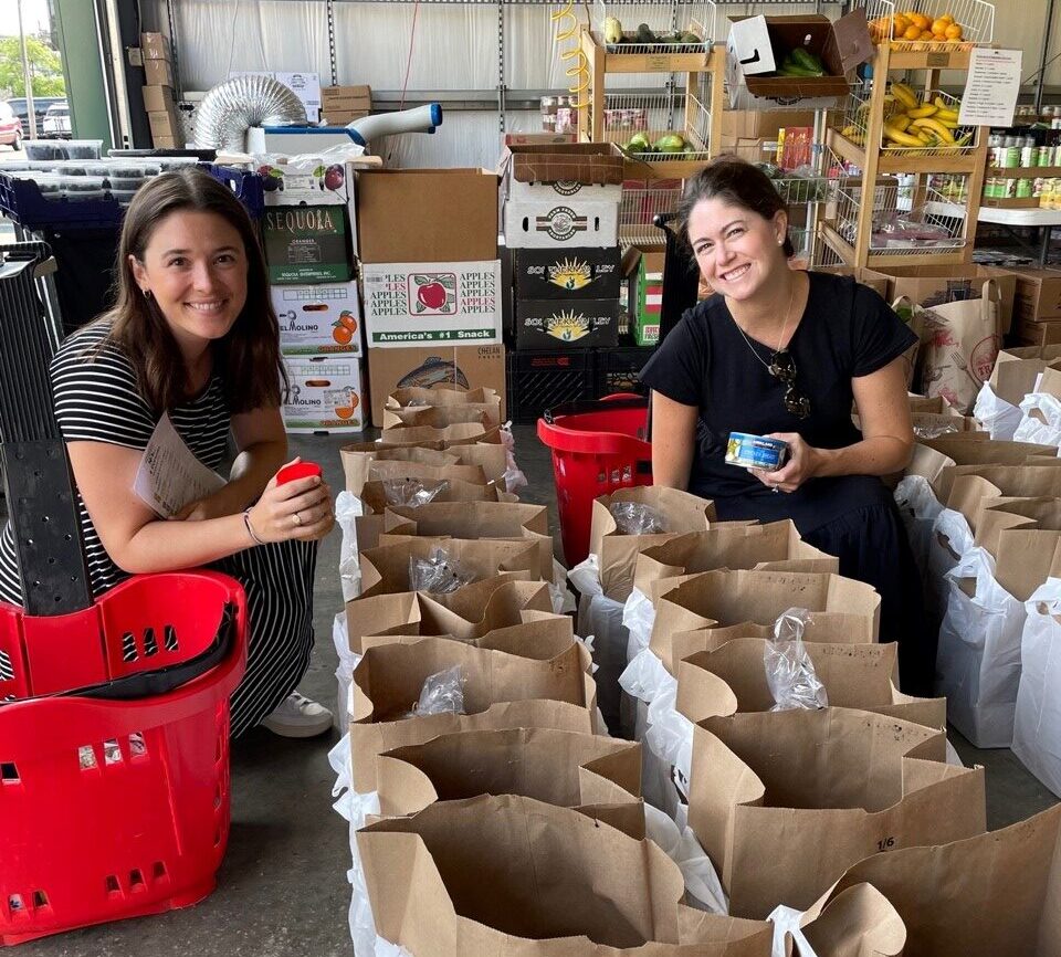 Two women kneel beside rows of filled paper grocery bags in a storage area, holding canned goods and smiling at the camera.