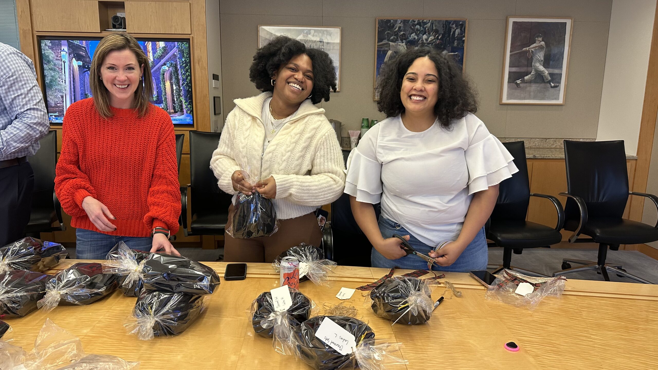 Three women standing behind a table with wrapped gifts, smiling and assembling items in an office setting.