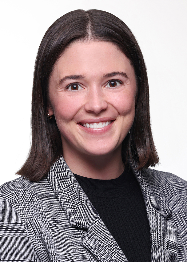 Woman with straight brown hair wearing a gray plaid blazer and black top, smiling at the camera against a plain white background.