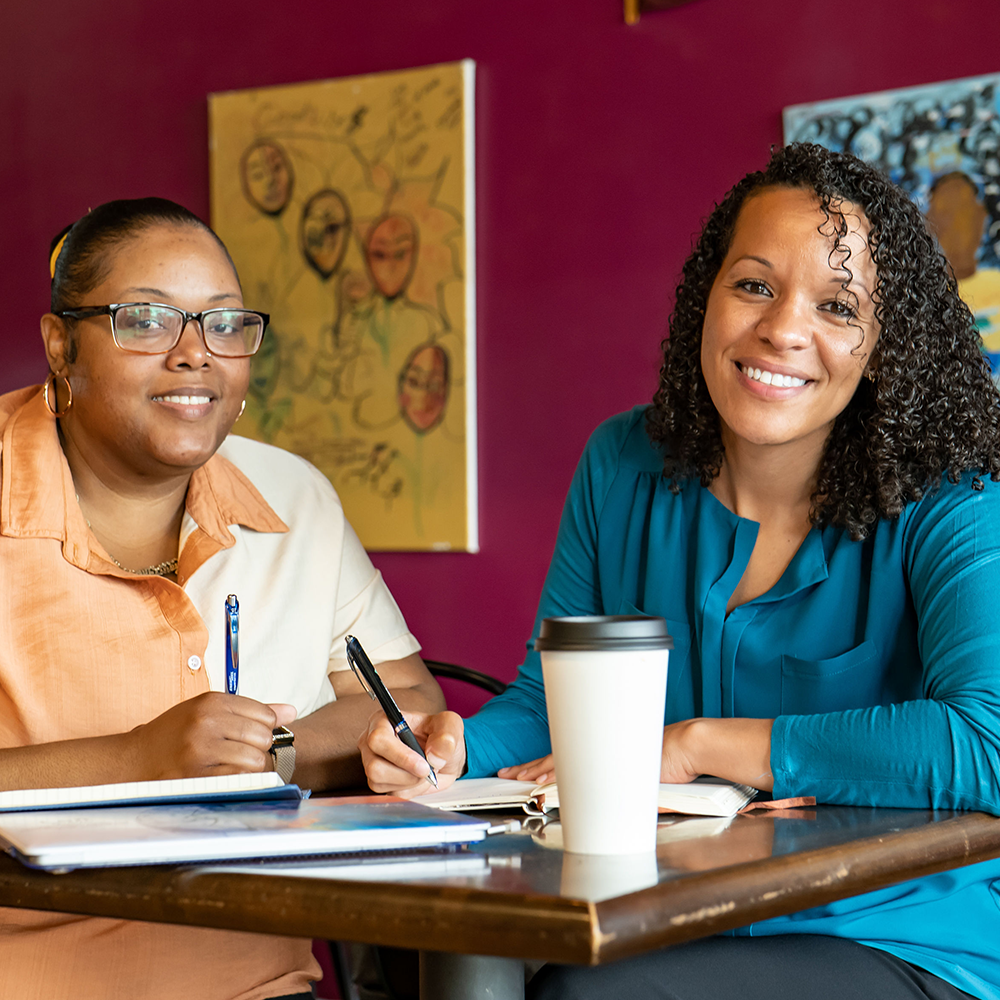Two women sit at a table with notebooks and pens, smiling at the camera during a Future Forward CT meeting. A coffee cup and colorful paintings are visible in the background.