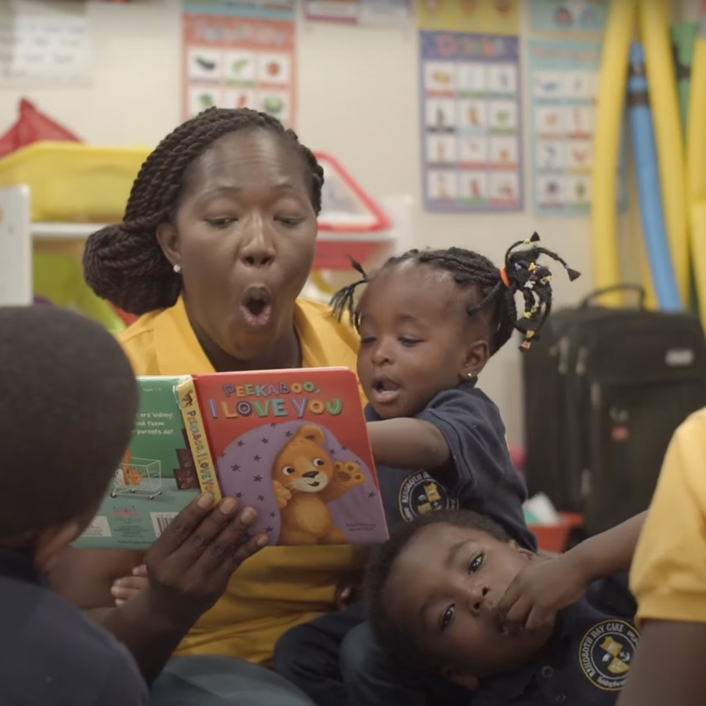 A woman reads a children's book to two young kids in a Future Forward CT classroom; one child points at the book while the other rests their head on her lap.