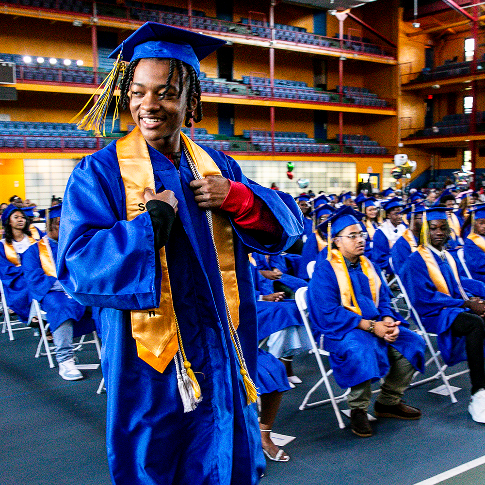 A student in a blue cap and gown walks past rows of seated graduates during the Future Forward CT commencement ceremony in a gymnasium.