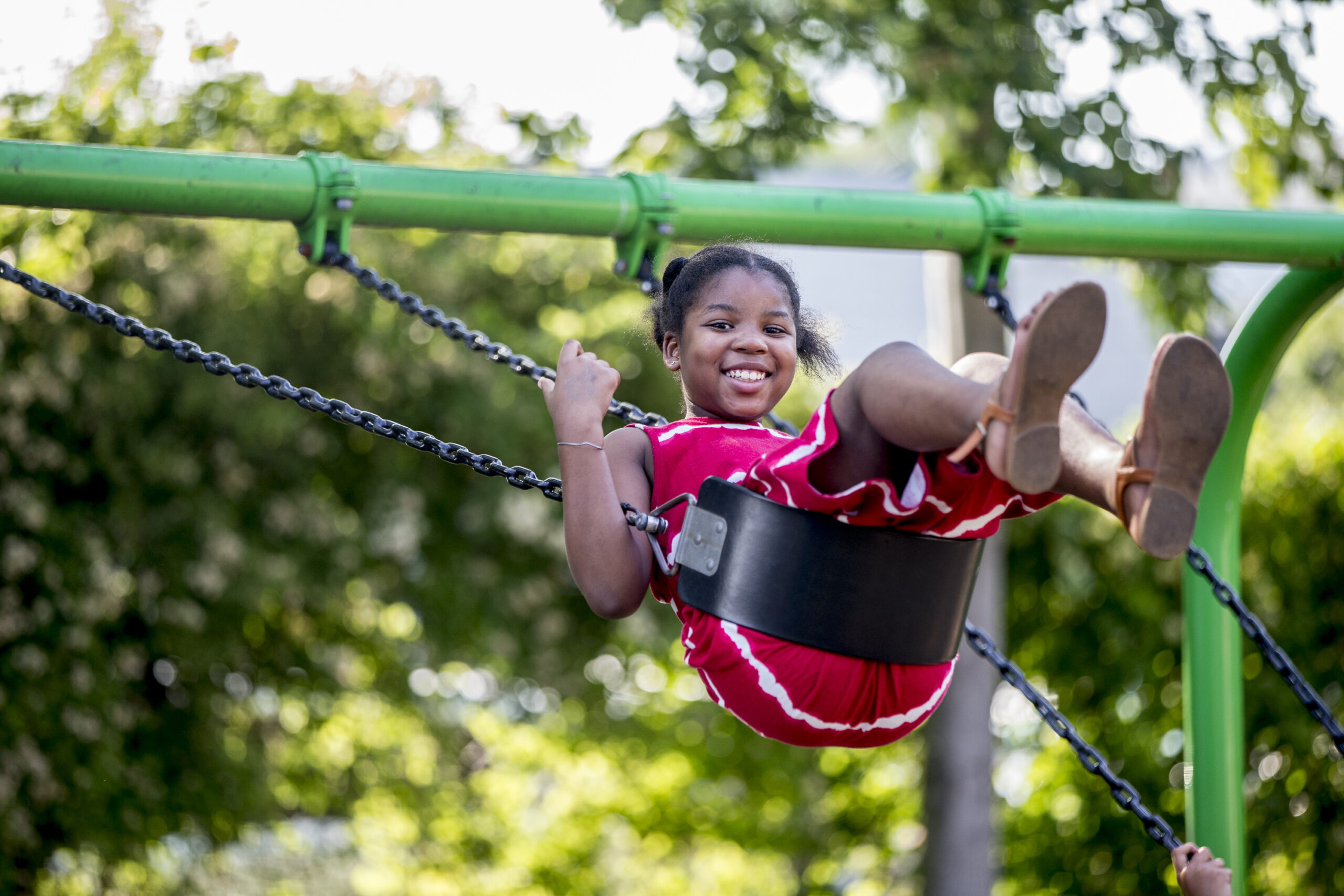 A young girl in a red dress smiles while swinging on a green playground swing set outdoors, capturing the joyful spirit of Who We Serve.
