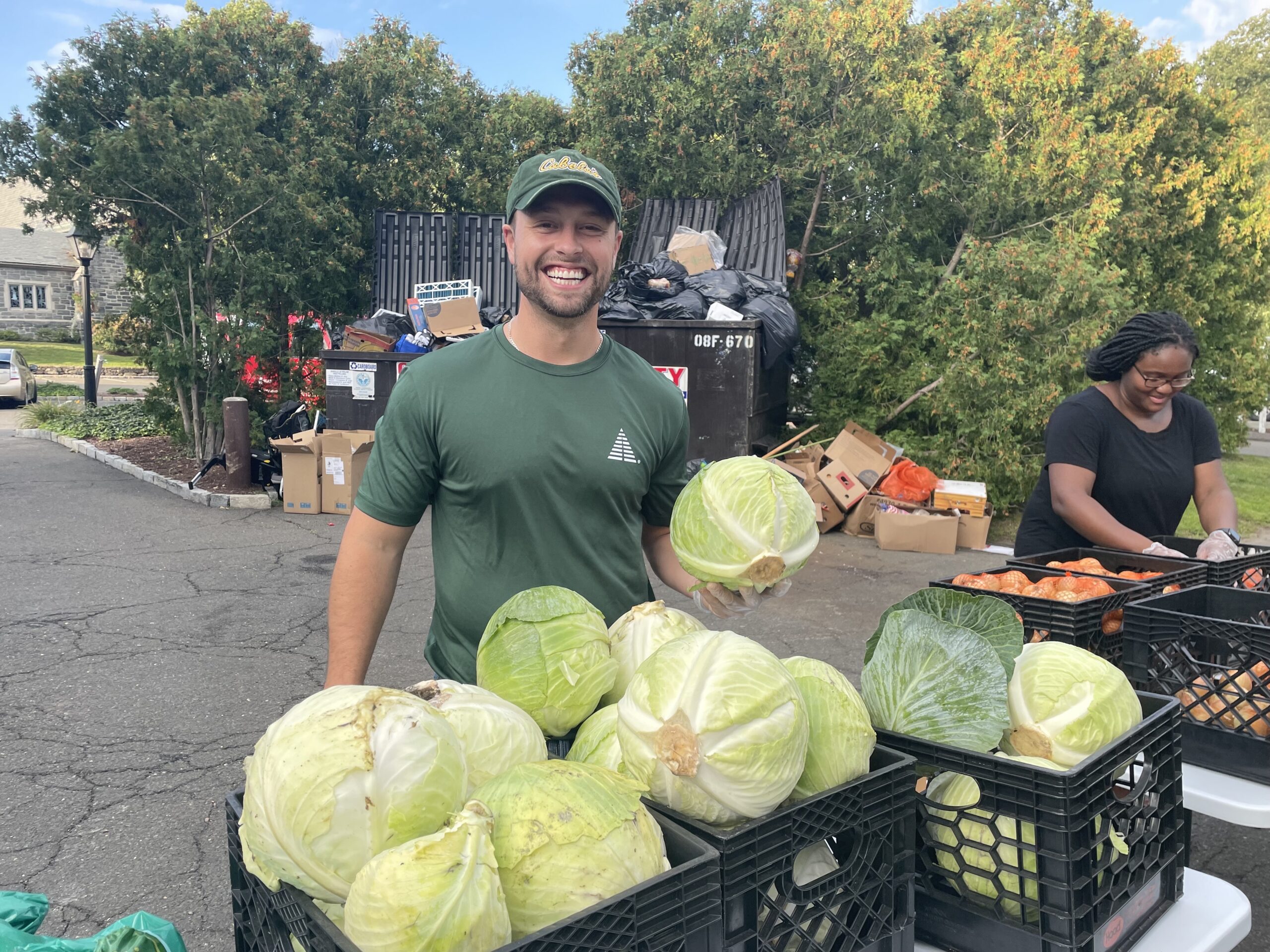 A man in a green shirt smiles while holding a cabbage at an outdoor market stall with crates of cabbages and other produce. Another person sorts vegetables nearby. Trees and dumpsters are in the background.